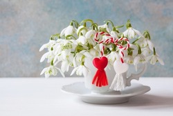 Postcard for the holiday of March 1. Snowdrops flowers in a cup on the table, colorful blue wall background and red and white martenitsa with hearts.