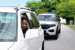 Young black man with a very worried look on his face is looking at the police car that has pulled him over in his rearview mirror reflecting the problems between citizens and some police departments.
