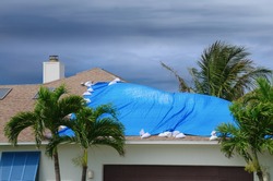 Storm damaged roof on house with a protective blue plastic tarp spread over hole in the shingles and rooftop.