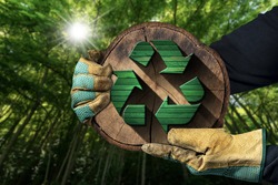 Hands with protective work gloves holding a recycling symbol made of green and brown wood inside of a cross section of a tree trunk. Sustainable Resources concept. Green forest on background.
