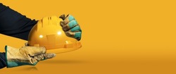 Manual worker with protective work gloves holding a yellow safety helmet on a yellow and orange background with copy space and reflections. Concept of workplace security.