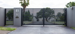 Metal driveway property entrance gates set in concrete fence with garden trees  in background