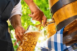 Bavarian man in apron is pouring a large lager beer in tap from wooden beer barrel in the beer garden. Background for Oktoberfest, folk or beer festival (German for: O’zapft is!) 
