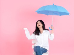 Happy Asian teenager girl holding blue umbrella and sitting on white chair, pink background.