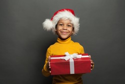 Black child Santa holding red Christmas box with white silky ribbon on gray background