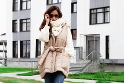 Fashionable woman dressed beige quilted gilet with belt, warm white knitted sweater, scarf, jean. Trend women's clothing for autumn or spring
