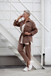 Fashionable woman stands. Short hair Model wears beige business suit with pants and jacket, belt purse, fashion accessories, loafers. Trend multilayered outfit. Fashion, style clothes