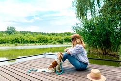Full length shot of smiling woman sitting next to her cute cavalier spaniel puppy on the pier while relaxing at the lake. 