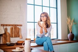 Middle aged woman sitting on the kitchen’s counter and drinking her morning tea while daydreaming. 