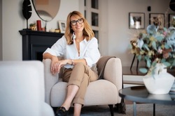 Portrait of attractive middle aged woman relaxing in an armchair at home. Blond haired female wearing eyeglasses and white shirt.