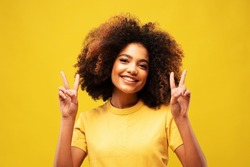 lifestyle, emotion and people concept: Photo of adorable shiny african woman wear yellow t-shirt showing v-sign smiling isolated yellow color background