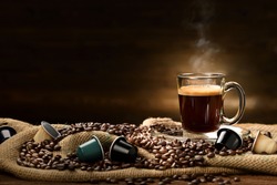 Cup glass of coffee with smoke and coffee beans and coffee capsules on burlap sack on old wooden background