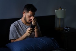 Young Asian man having sore and tired eyes when using smartphone while lying in bed at night. Young man cannot sleep from insomnia	