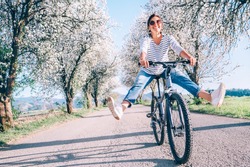 Happy smiling woman cheerfully spreads legs on bicycle on the country road under blossom trees. Spring is comming concept image.