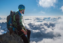 Bearded Climber in a safety harness, helmet, and on body wrapped climbing rope with sitting at 3600m altitude on a cliff and looking at  picturesque clouds during Mont Blanc ascending, France route