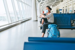 Lonely female solo traveler with backpack sitting in the empty airport passenger transfer hall in protective face mask and looking out large windows. Traveling in worldwide pandemic time concept image