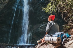 Smiling Woman with backpack dressed in active trekking clothes sitting near the mountain river waterfall and enjoying the splashing Nature power. Traveling, trekking and nature concept image.