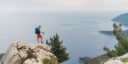 Young hiker backpacker man using trekking poles enjoying the Mediterranean Sea on clifftop during Lycian Way trekking walk. Famous Likya Yolu Turkish route near Letoon. Active vacations concept image 