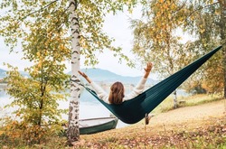 The young woman cheerfully rose arms up while she swinging in a hammock between the birch trees on the mountain lake bank. Out-of-town Outdoor Recreation in Nature concept image.