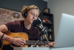 Home sound studio young teenager portrait playing guitar in Headphones recording voice music using a microphone and laptop computer in the kids room. Modern audio recording technology concept image.