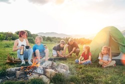 Group of smiling kids has a merry conversation near a smoky campfire. They drinking tea from a thermos, two brothers set up the green tent. Happy family outdoor picnic camping activities concept