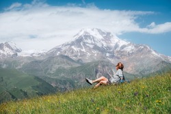 Backpacker woman sitting on a green grass hill and enjoying snowy slopes of Kazbek 5054m mountain with a backpack while she walking by green grass hill. East Caucasus mountains, Georgia.