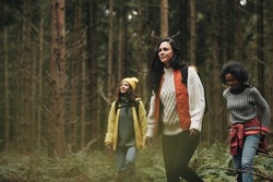 Three diverse young women in outdoor gear walking on a trail together through a dense forest