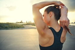 Athletic woman warming up before a workout standing facing the early morning rising sun on a rural road doing stretching exercises, close up with copy space