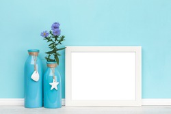 White blank horizontal picture frame, decorated vases with blue daisy flowers in front of aqua colored wall, Poster mockup, blank area isolated with clipping path