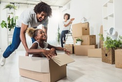 Young Smiling Happy African-american Family Unpacking During Move. New Home. Little preschooler daughter sitting in cardboard box, father rolling her. Smiling mother looking them.