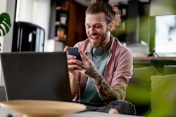 Young cheerful man with a sleeve tattoo using laptop computer and smartphone. Freelance entrepreneur working from home using banking apps.