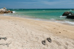 Flip flops on sand beach with stone arch and turquoise sea against blue sky in Ko Man Klang, Rayong, Thailand.