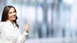 Portrait of happy smiling young excited businesswoman, showing thumb up hand sign gesture. Success in business concept studio shot. Standing against blurred office background.