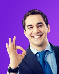 Excited cheerful smiling senior businessman in blue suit and tie, showing okay ok hand sign, over violet purple color background. Happy confident man gesturing. Business success concept.