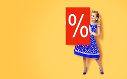 Beautiful woman in pinup dress holding, showing red board with procents % sign. Full body pin up girl with advertising offer, isolated on yellow color background. Sales, discounts rebates ad concept.