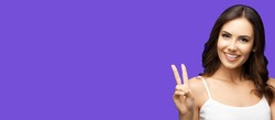 Photo of woman showing two fingers or victory hand sign gesture, on violet purple studio background. Happy smiling gesturing brunette girl. Wide banner composition with mock up copy space.