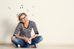 Portrait of happy female listening musical composition in earphones with sheet music and clef drawn on the wall. Young pretty Caucasian woman sitting on wooden floor in her house. 