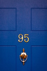 House number 95 on a blue wooden front door with golden digits and knocker with integrated peephole