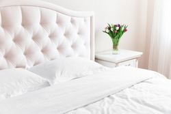 Bedroom with bed, white bedding, and bedside table with bouquet of tulips in a vase. White pillows, duvet and duvet case on bed with beige headboard.  Bed with clean white pillows and bed sheets.