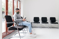 Young man with face mask sitting in a waiting room of a hospital or office looking at smartphone - focus on the man