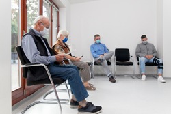 Senior couple with face masks sitting in a waiting room of a hospital together with a young and mature man - focus on the old man in the foreground