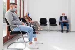 Young and old people with face masks keeping social distance in a waiting room of a hospital or office -  focus on the young man in the foreground