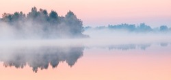 Dawn over the foggy lake. Beautiful dreamy view. Pink sky just before the sunrise and fog over water and trees with reflections on the river bank.