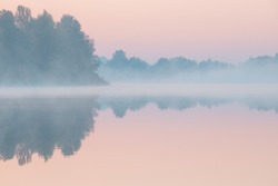 Dawn over the foggy lake. Beautiful dreamy view. Pink sky just before the sunrise and fog over water and trees with reflections on the river bank.