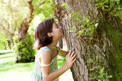 Child girl hugging kissing mature textured tree trunk in sunny forest park, smiling with eyes shut, love nature. Kid caring and protecting organic eco climate change resources. Healthy wellness planet