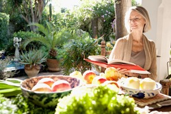Active mature attractive woman sitting in her home garden reading a cook book next to a table full of organic vegetables and fruit in abundance, during a sunny day outdoors.