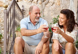 Portrait of a senior man and woman sitting on the stone steps of luxury hotel garden on holiday drinking wine and toasting in celebration, relaxing on vacation. Mature people, outdoors lifestyle.