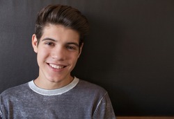 Close up portrait of a young hispanic teenager man looking at camera with a joyful smiling expression, against a black background at home, interior. Teenager being confident and smart, indoors.