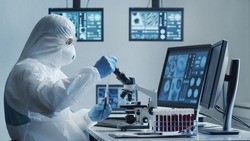 Scientist in protection suit and masks working in research lab using laboratory equipment: microscopes, test tubes. Coronavirus 2019-ncov hazard, pharmaceutical discovery, bacteriology and virology.