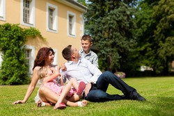 Young happy family sitting in the sun on the lawn in front of their new home - a villa